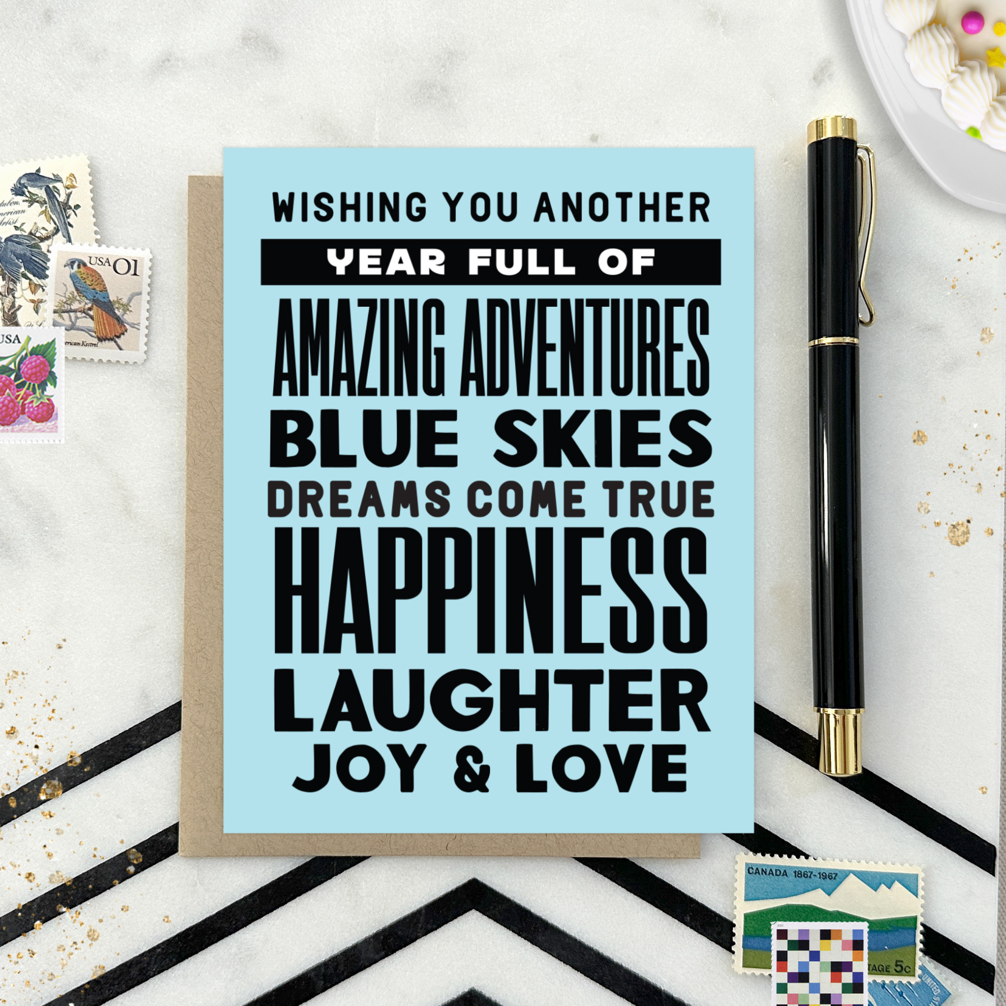 A birthday greeting card with blue background and black text, featuring stacked words and phrases like 'amazing adventures', 'blue skies', 'dreams come true', 'happiness', 'laughter', 'joy & love'
