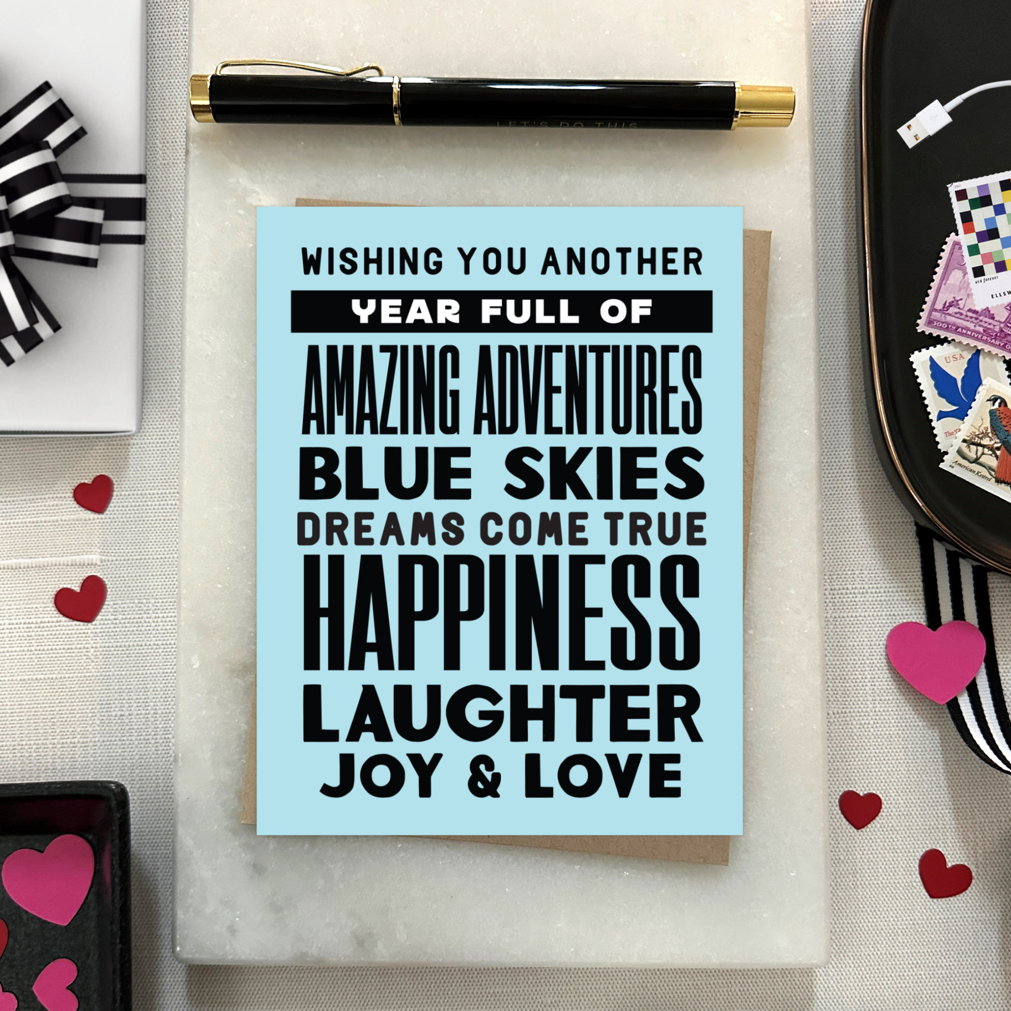 A birthday greeting card with blue background and black text, featuring stacked words and phrases like 'amazing adventures', 'blue skies', 'dreams come true', 'happiness', 'laughter', 'joy & love'
