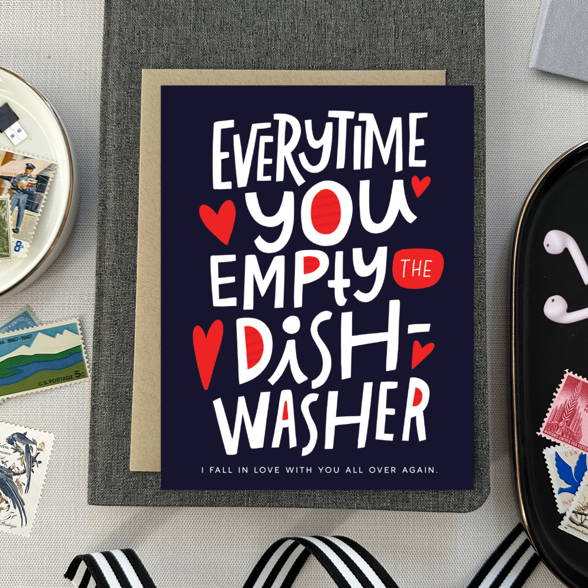 A Valentine's day greeting card with hand lettering that reads 'Everytime you empty the dishwasher I fall in love with you all over again.' It features red and red-hearts-shaped text on a dark background.