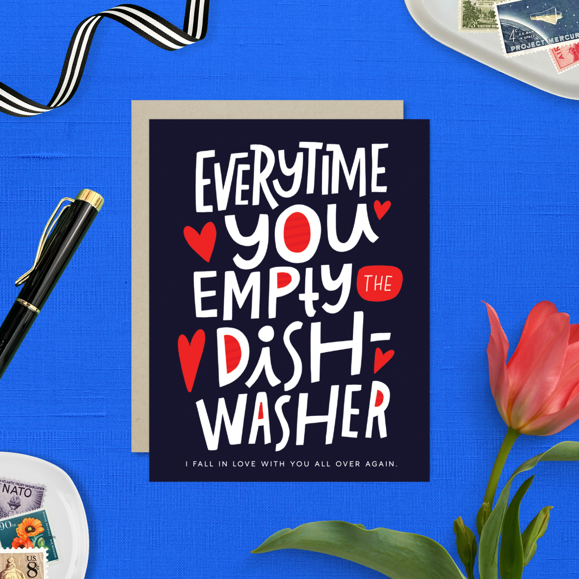 A Valentine's day greeting card with hand lettering that reads 'Everytime you empty the dishwasher I fall in love with you all over again.' It features red and red-hearts-shaped text on a dark background.