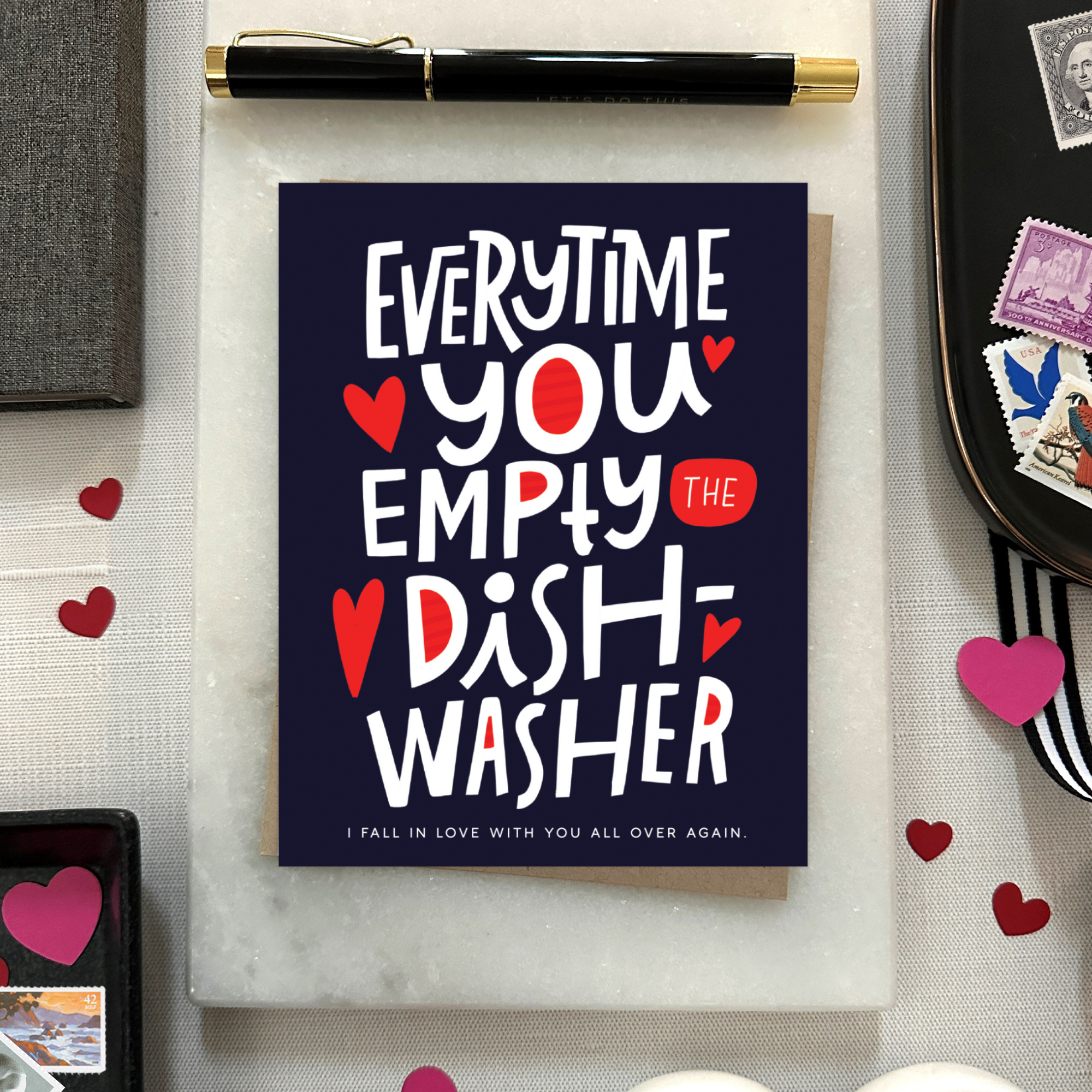 A Valentine's day greeting card with hand lettering that reads 'Everytime you empty the dishwasher I fall in love with you all over again.' It features red and red-hearts-shaped text on a dark background.
