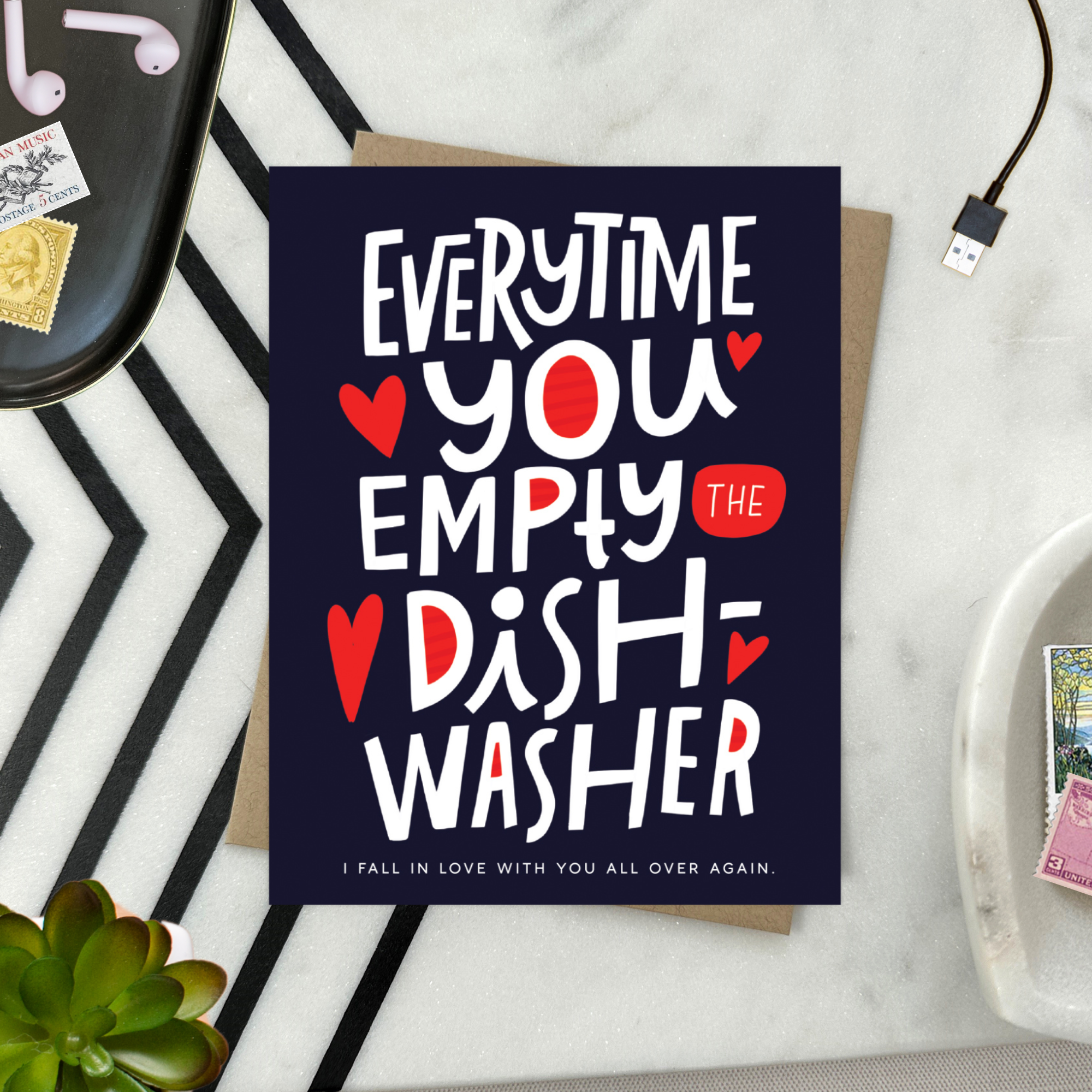 A Valentine's day greeting card with hand lettering that reads 'Everytime you empty the dishwasher I fall in love with you all over again.' It features red and red-hearts-shaped text on a dark background.