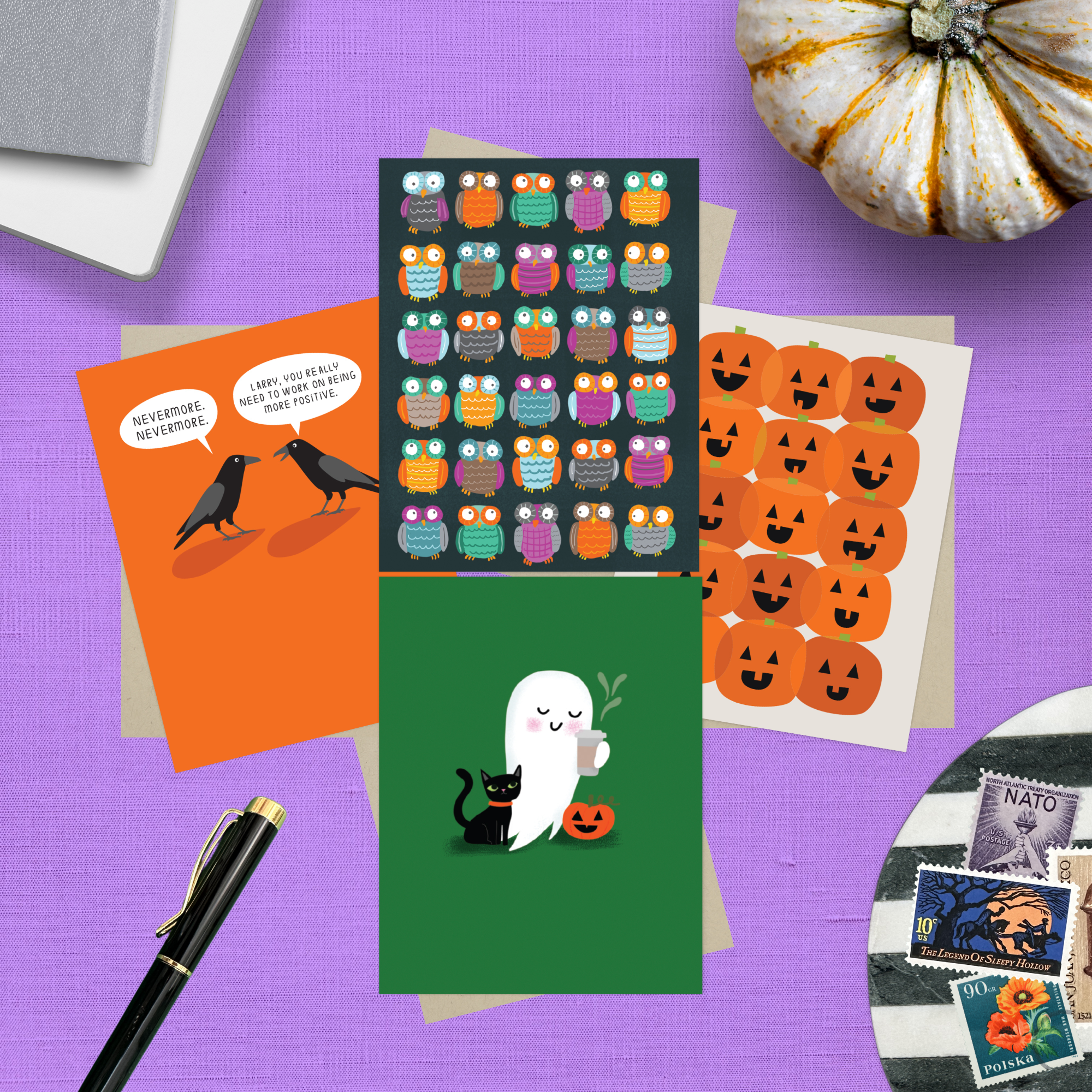 Collection of Halloween-themed greeting cards featuring an adorable ghost with black cat and coffee, one with 2 funny ravens saying "Nevermore," one with a jack-o-lantern pattern, and one with an owl pattern with stamps on a purple surface with a pumpkin and pen.