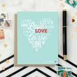 A heart-shaped Valentine's Day greeting card with the word 'love' arranged in a heart pattern using different fonts on a teal background, with a brown envelope.