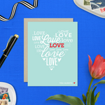 A heart-shaped Valentine's Day greeting card with the word 'love' arranged in a heart pattern using different fonts on a teal background, with a brown envelope.