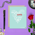 A heart-shaped Valentine's Day greeting card with the word 'love' arranged in a heart pattern using different fonts on a teal background, with a brown envelope.