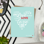 A heart-shaped Valentine's Day greeting card with the word 'love' arranged in a heart pattern using different fonts on a teal background, with a brown envelope.