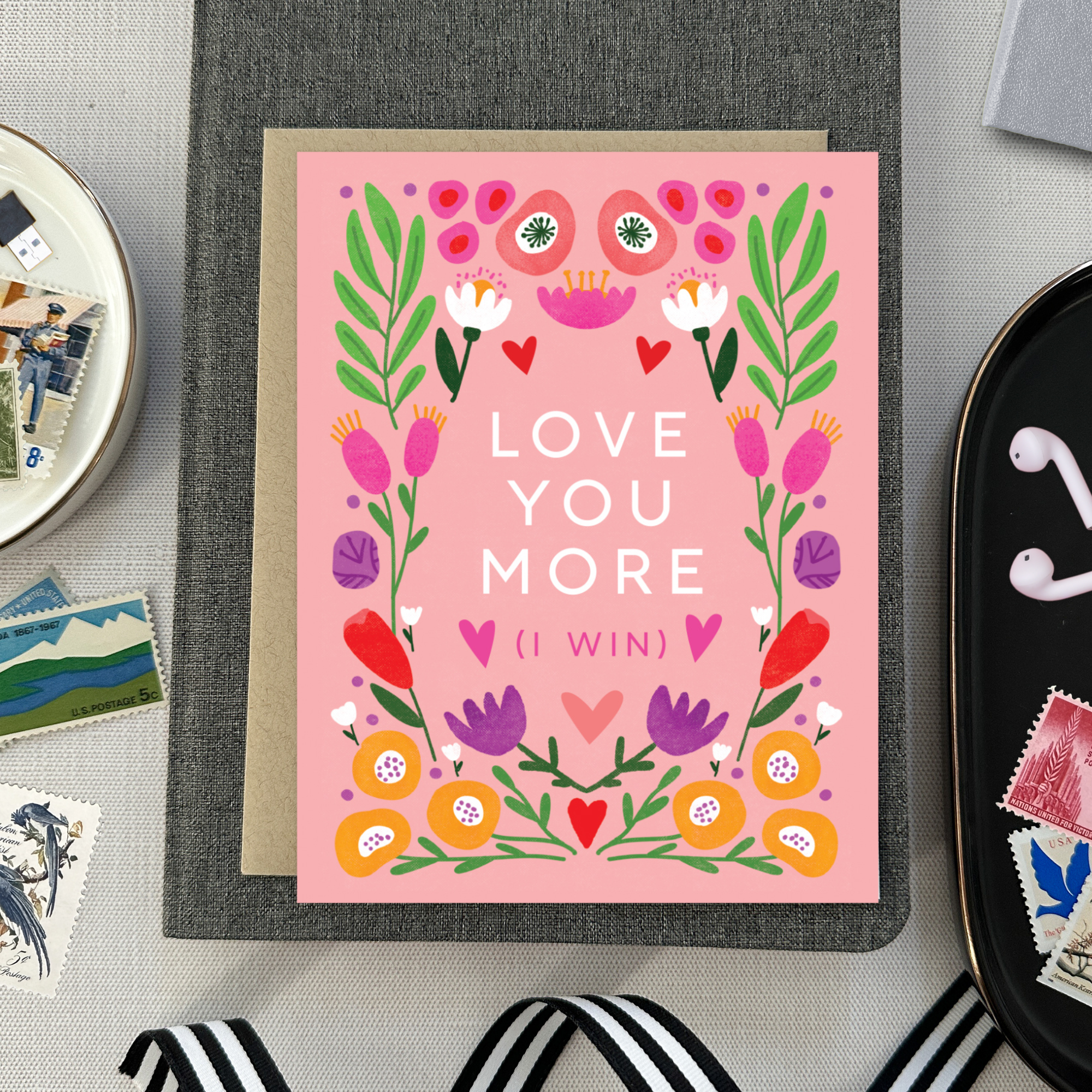 A Valentine's Day greeting card with a bold, symmetric botanical design and the text 'LOVE YOU MORE (I WIN)' surrounded by colorful flowers and hearts on a pink background.