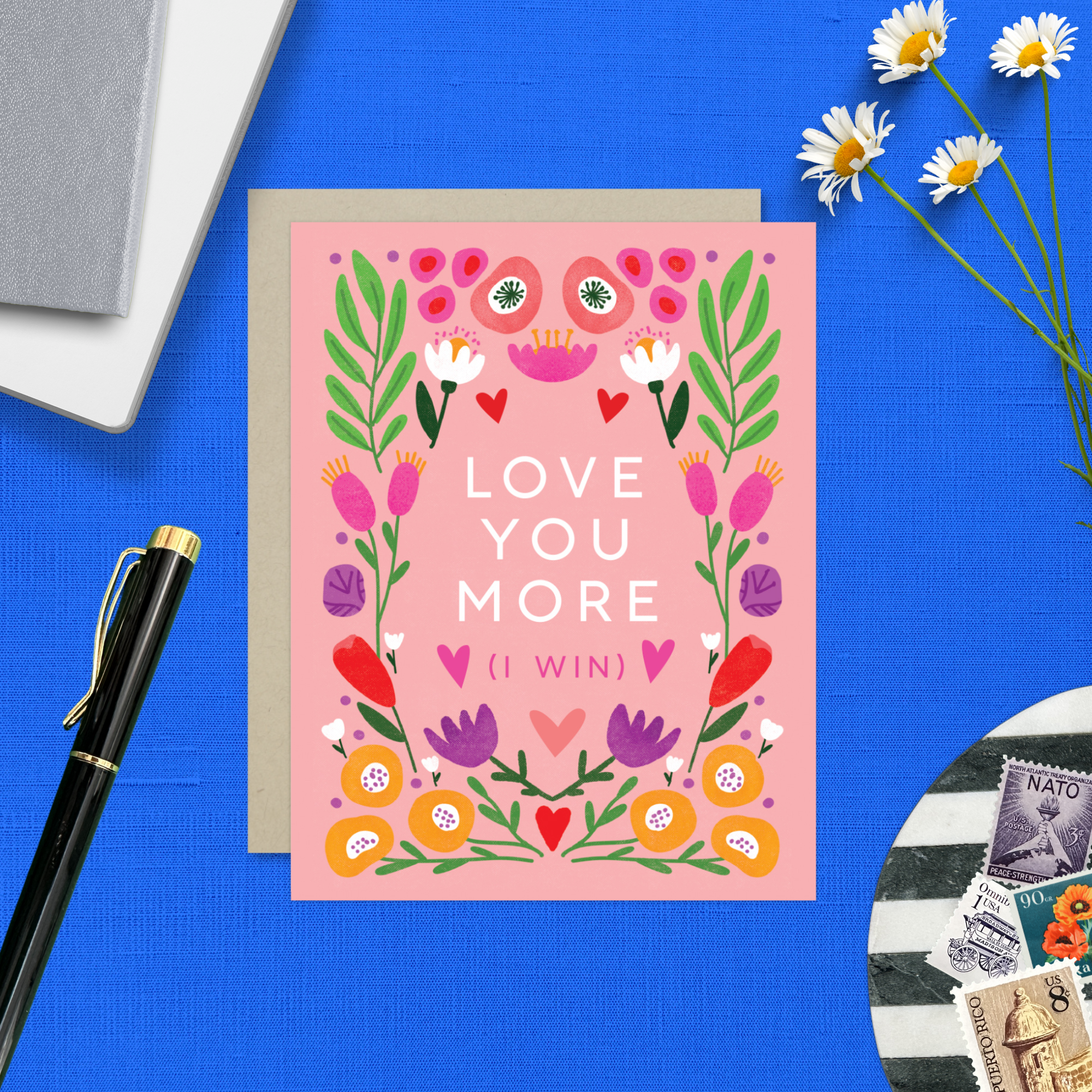 A Valentine's Day greeting card with a bold, symmetric botanical design and the text 'LOVE YOU MORE (I WIN)' surrounded by colorful flowers and hearts on a pink background.
