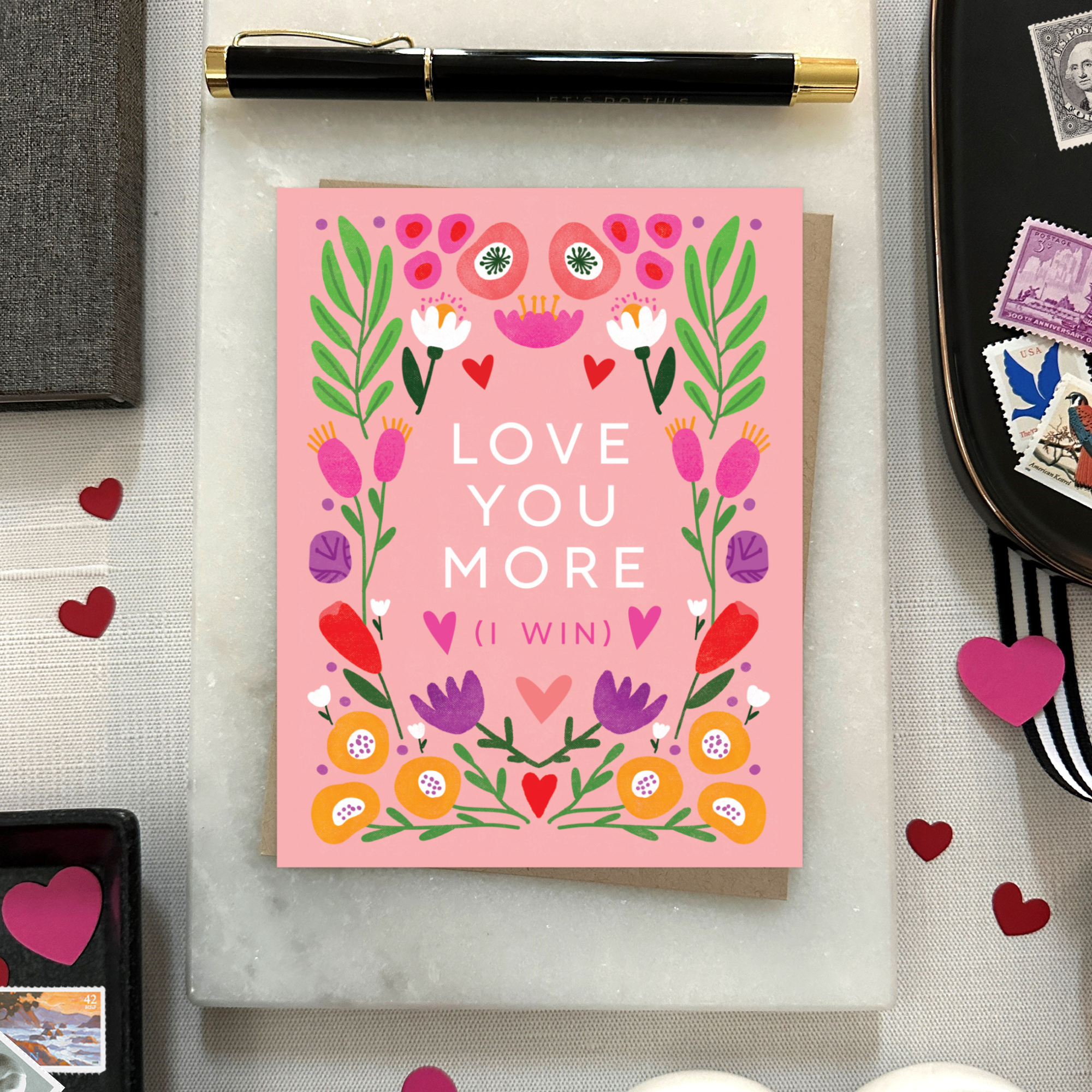 A Valentine's Day greeting card with a bold, symmetric botanical design and the text 'LOVE YOU MORE (I WIN)' surrounded by colorful flowers and hearts on a pink background.