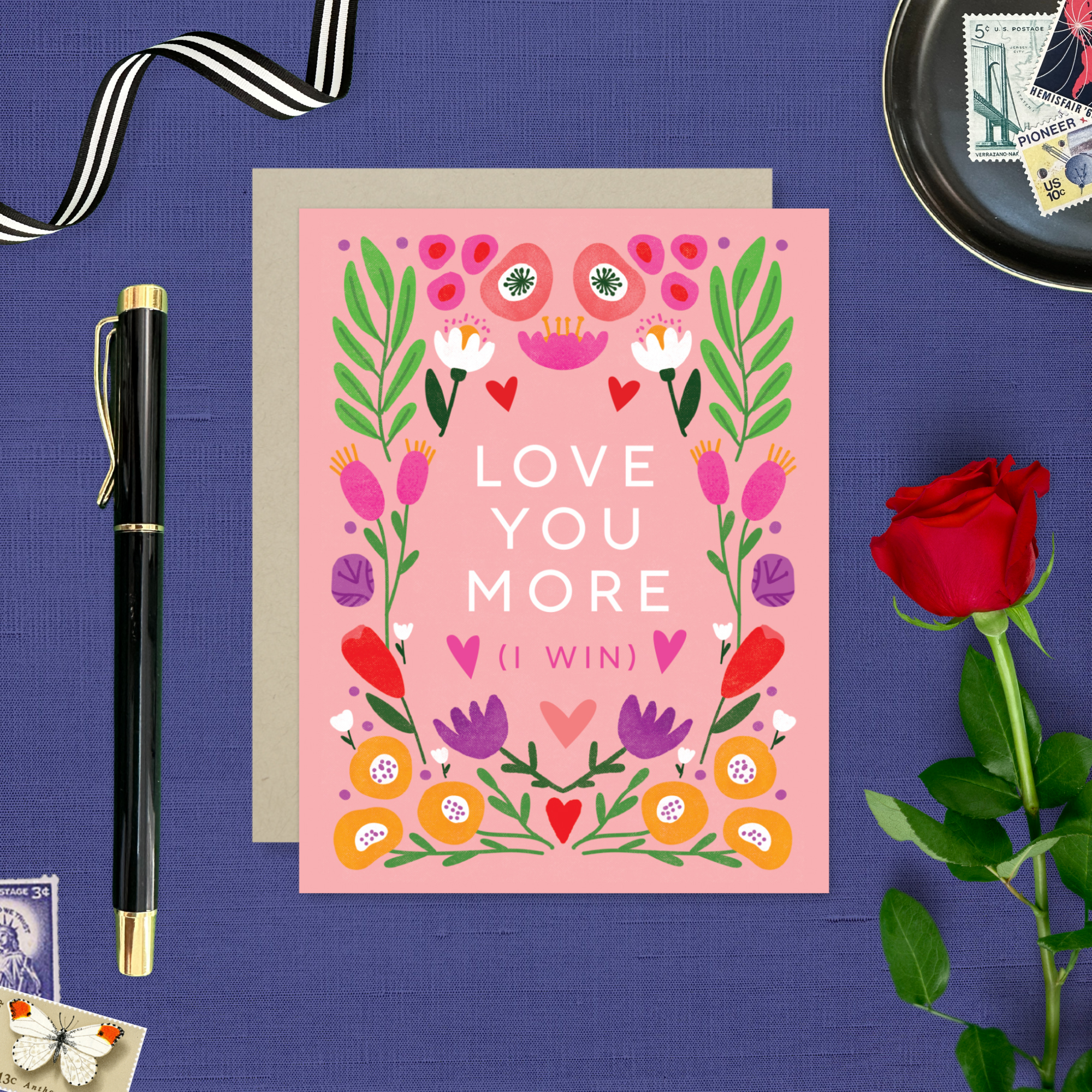 A Valentine's Day greeting card with a bold, symmetric botanical design and the text 'LOVE YOU MORE (I WIN)' surrounded by colorful flowers and hearts on a pink background.