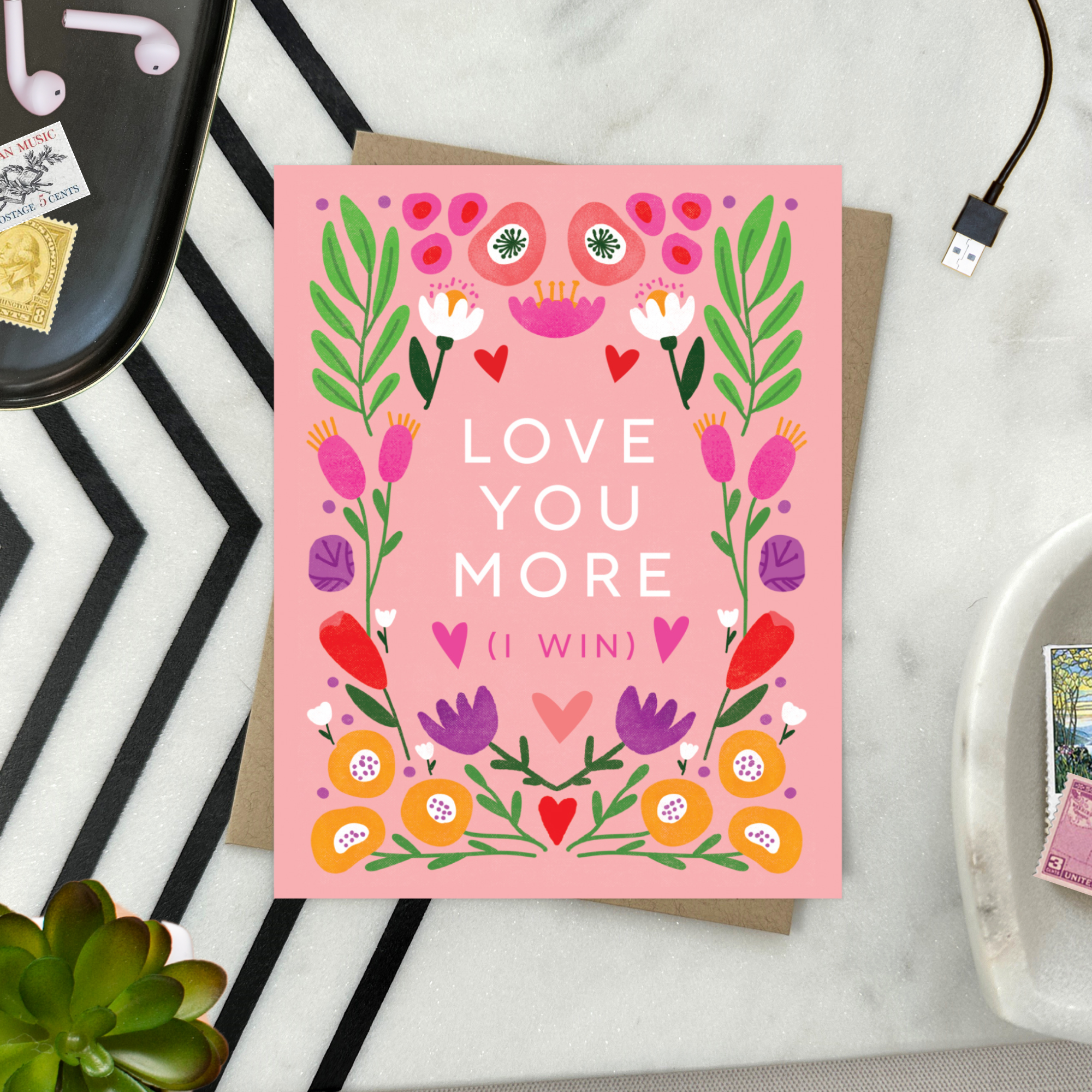 A Valentine's Day greeting card with a bold, symmetric botanical design and the text 'LOVE YOU MORE (I WIN)' surrounded by colorful flowers and hearts on a pink background.
