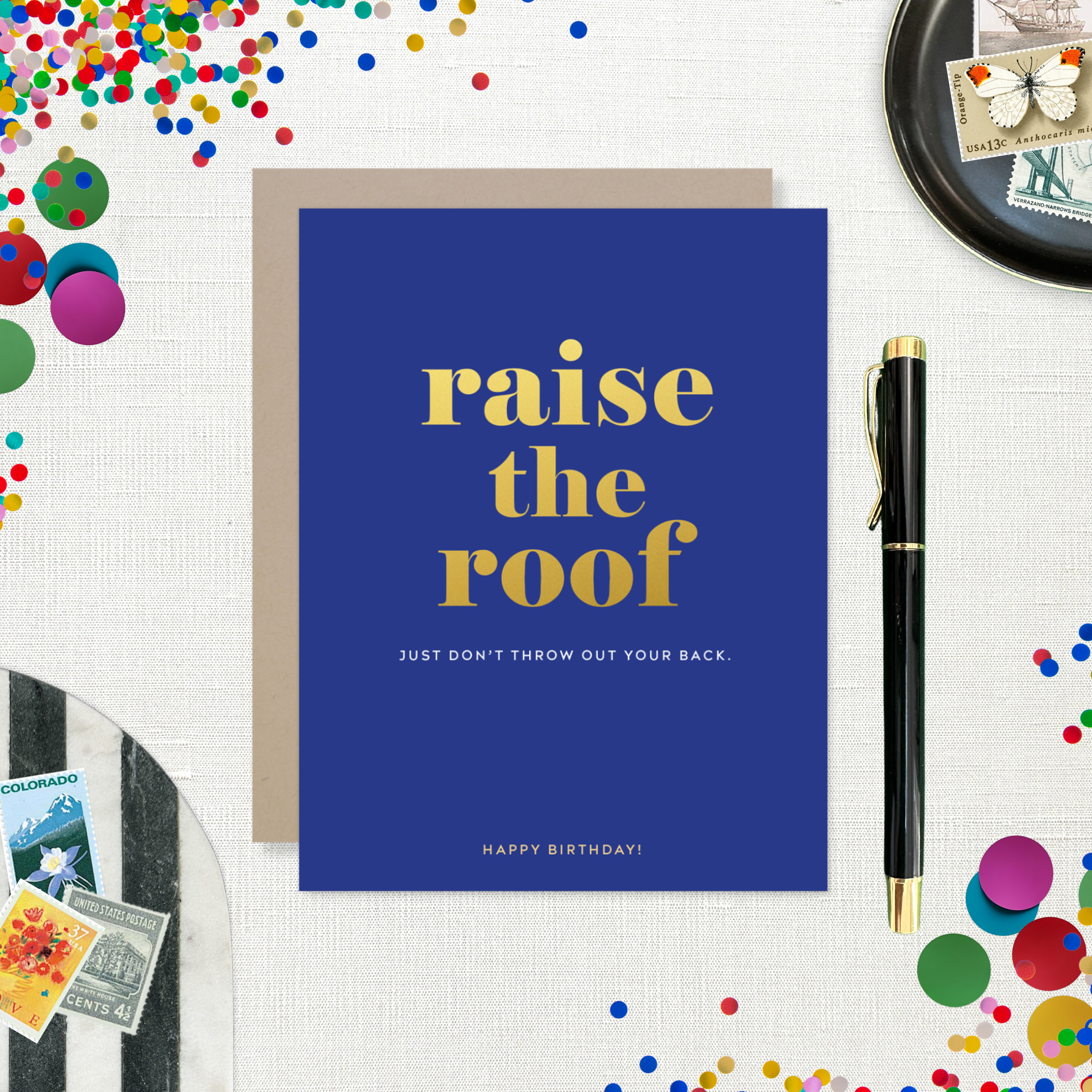 A birthday greeting card with 'raise the roof' printed in gold foil. Below it, in smaller gold foil text, reads 'Just don’t throw out your back.