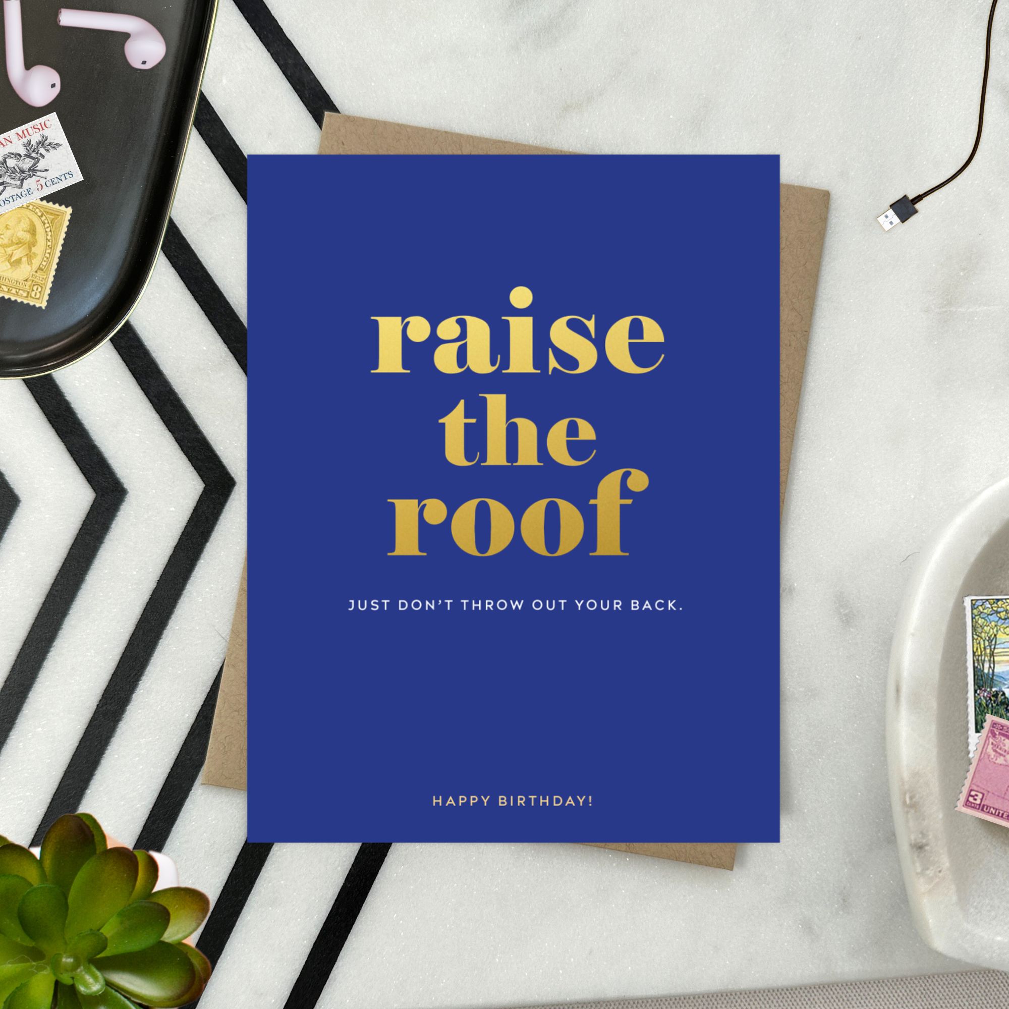 A birthday greeting card with 'raise the roof' printed in gold foil. Below it, in smaller gold foil text, reads 'Just don’t throw out your back.