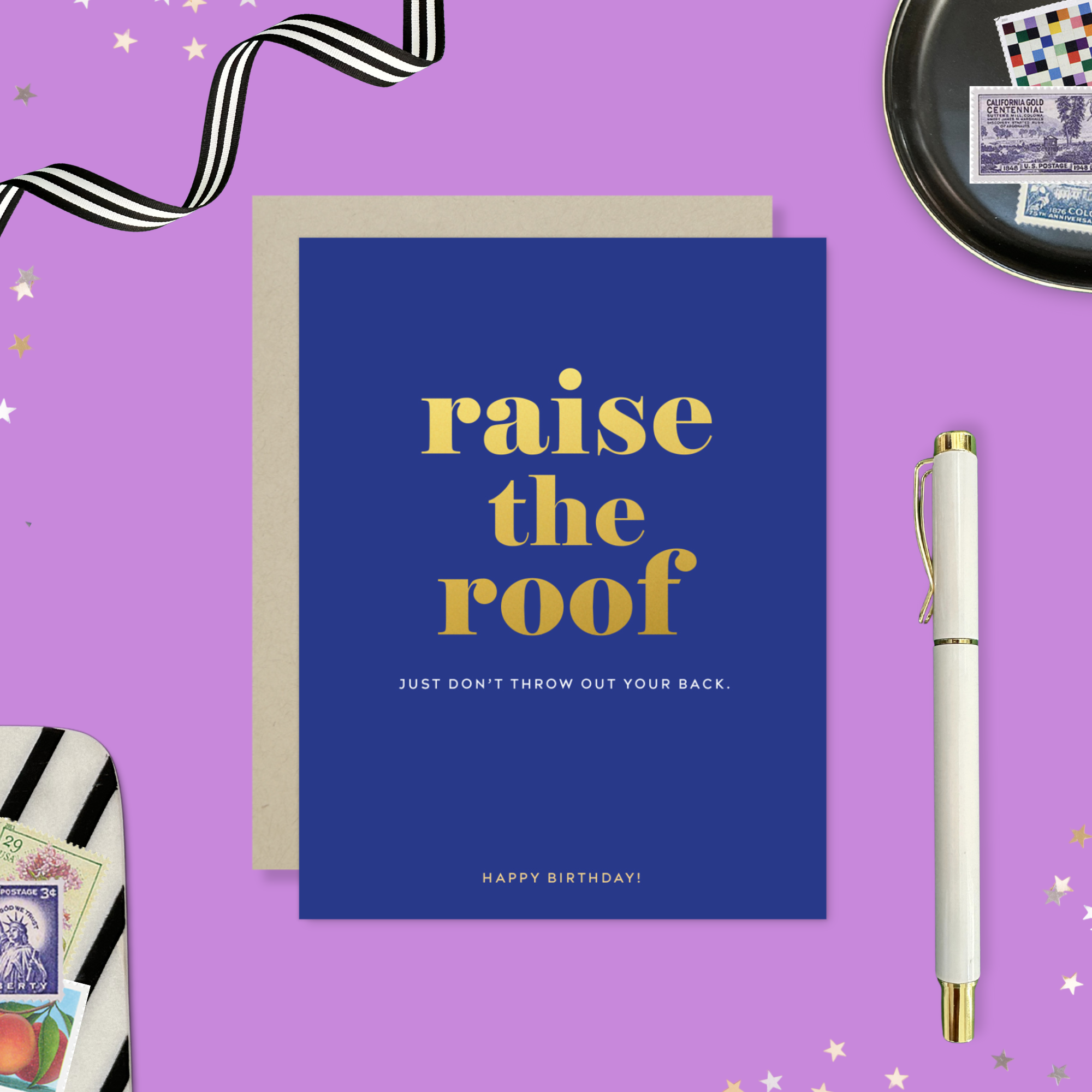 A birthday greeting card with 'raise the roof' printed in gold foil. Below it, in smaller gold foil text, reads 'Just don’t throw out your back.