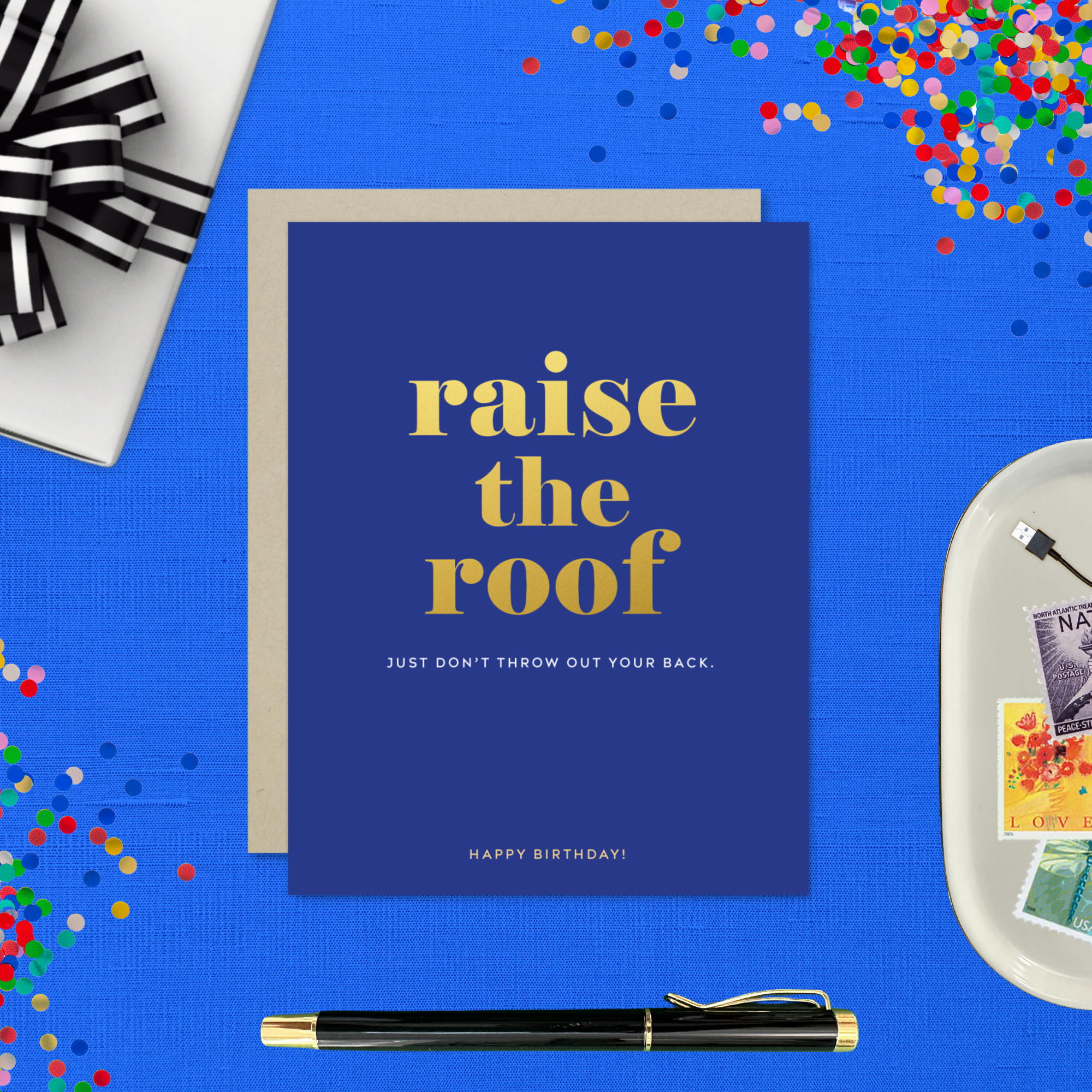 A birthday greeting card with 'raise the roof' printed in gold foil. Below it, in smaller gold foil text, reads 'Just don’t throw out your back.