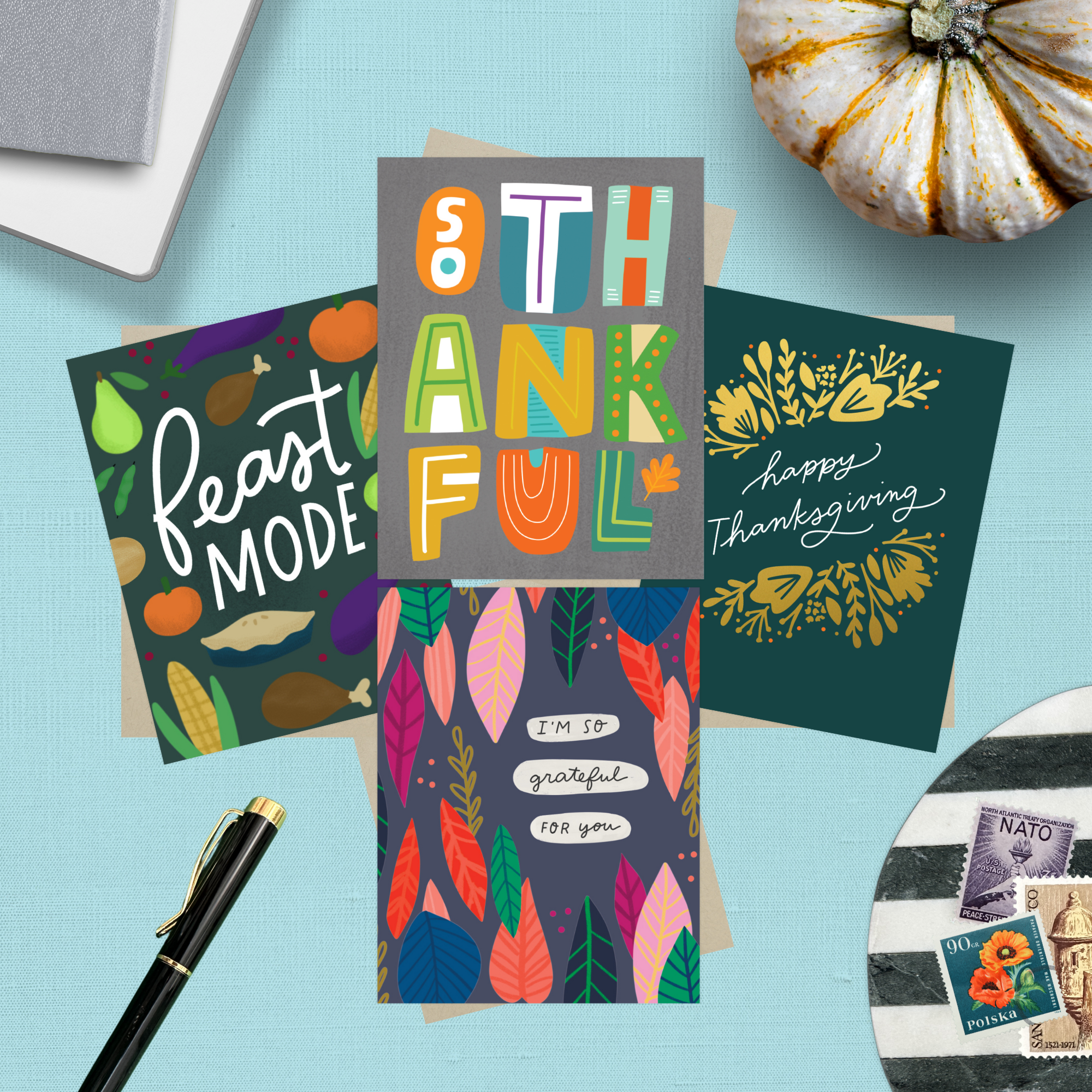 Collection of Thanksgiving-themed cards on a blue background with a pen and decorative items featuring "I'm so grateful for you," "Happy Thanksgiving," "Feast Mode," and "So Thankful."