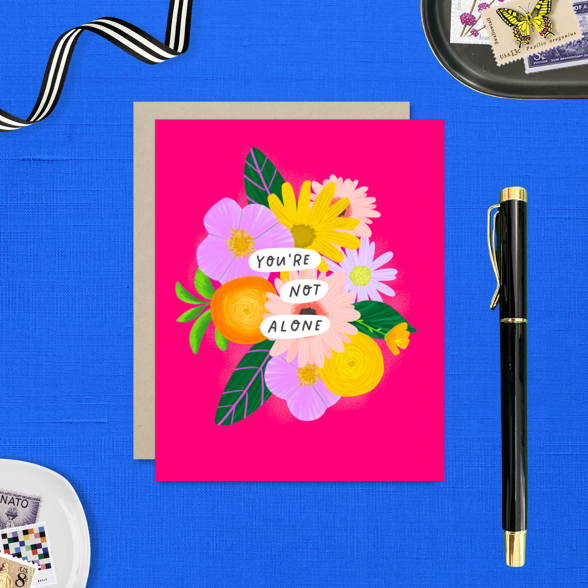 Colorful friendship or encouragement greeting card featuring a modern floral bouquet in pinks, oranges, yellows, and lavenders with the handwritten words, "You Are Not Alone" on a hot pink background with a matching kraft envelope.