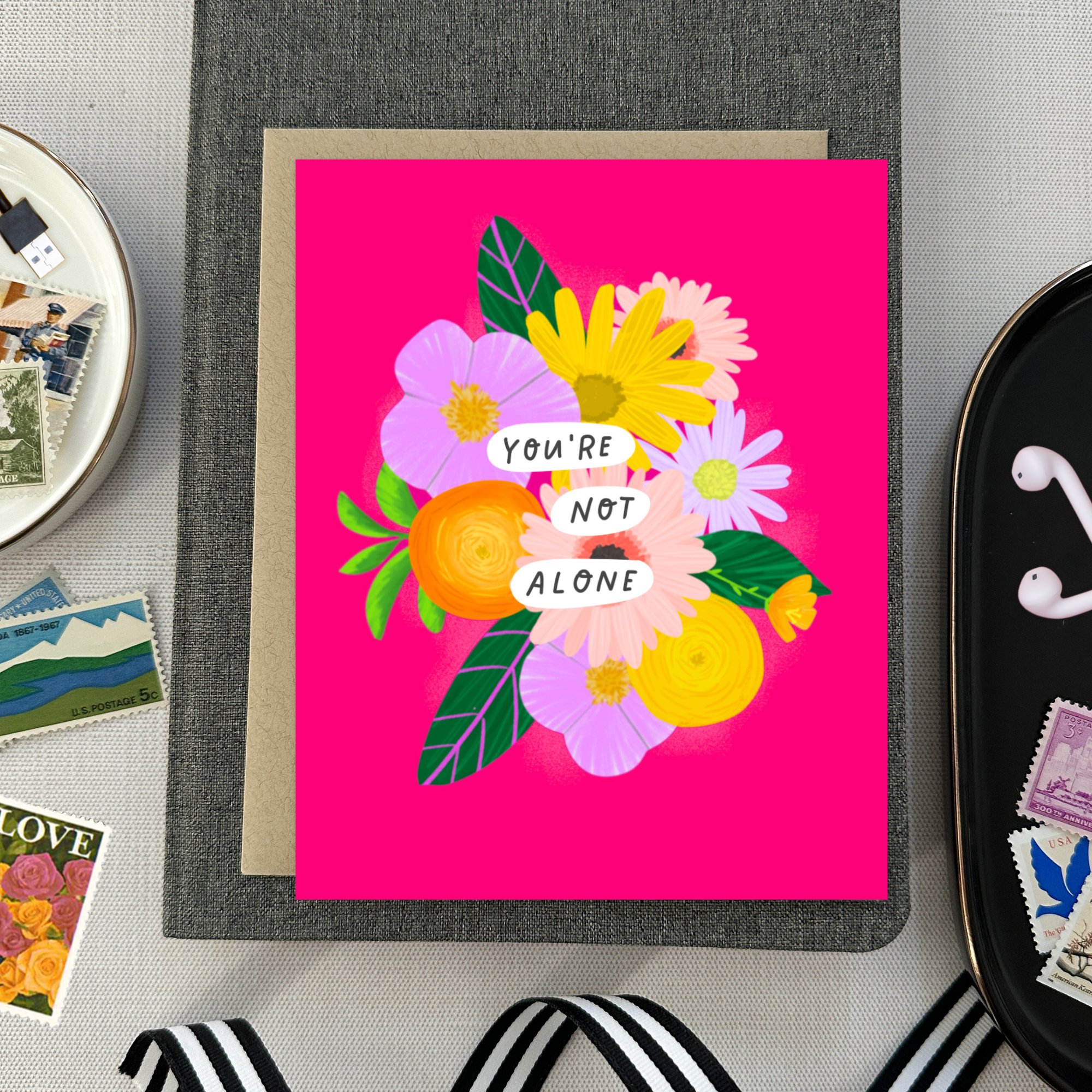 Colorful friendship or encouragement greeting card featuring a modern floral bouquet in pinks, oranges, yellows, and lavenders with the handwritten words, "You Are Not Alone" on a hot pink background. Includes a matching kraft envelope.