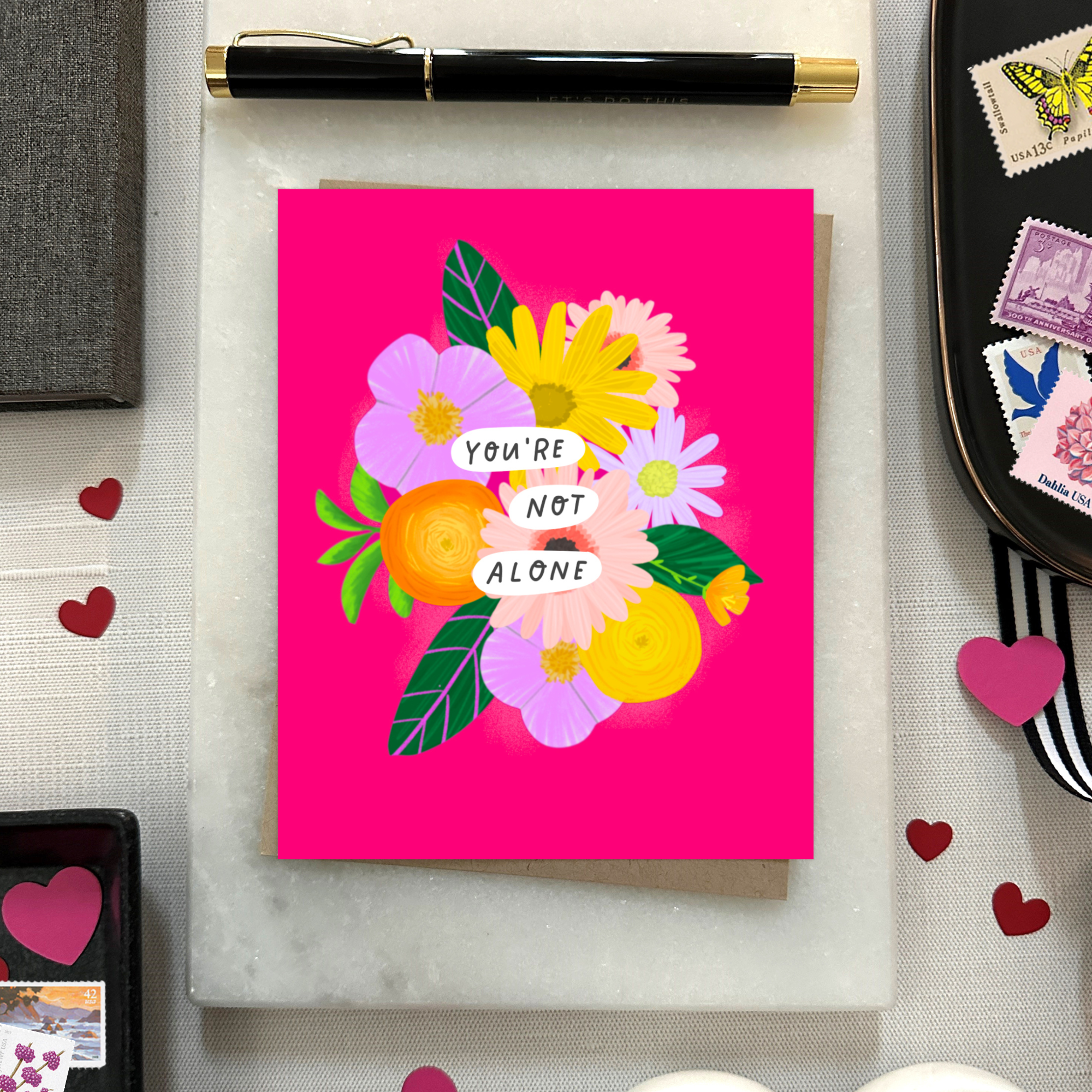 Colorful encouragement or friendship greeting card featuring a modern floral bouquet in pinks, oranges, yellows, and lavenders with the handwritten words, "You Are Not Alone" on a hot pink background with included matching kraft envelope.