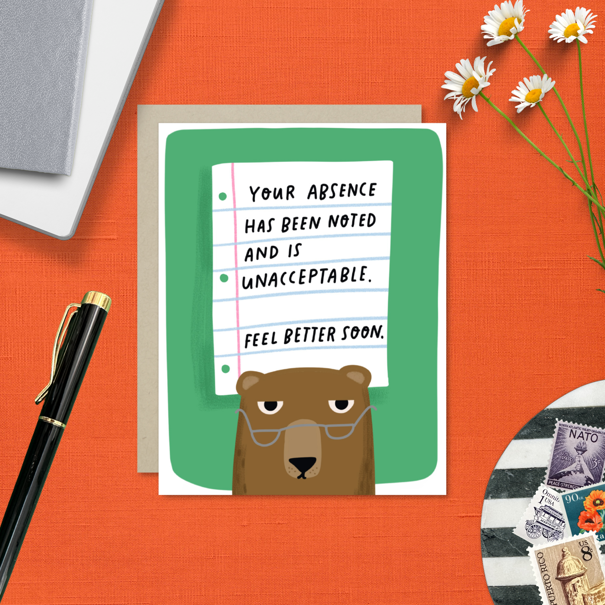 Get well Greeting card with bear illustration and text on an orange surface with a pen and flowers.