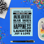 A birthday greeting card with blue background and black text, featuring stacked words and phrases like 'amazing adventures', 'blue skies', 'dreams come true', 'happiness', 'laughter', 'joy & love'