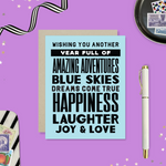 A birthday greeting card with blue background and black text, featuring stacked words and phrases like 'amazing adventures', 'blue skies', 'dreams come true', 'happiness', 'laughter', 'joy & love'