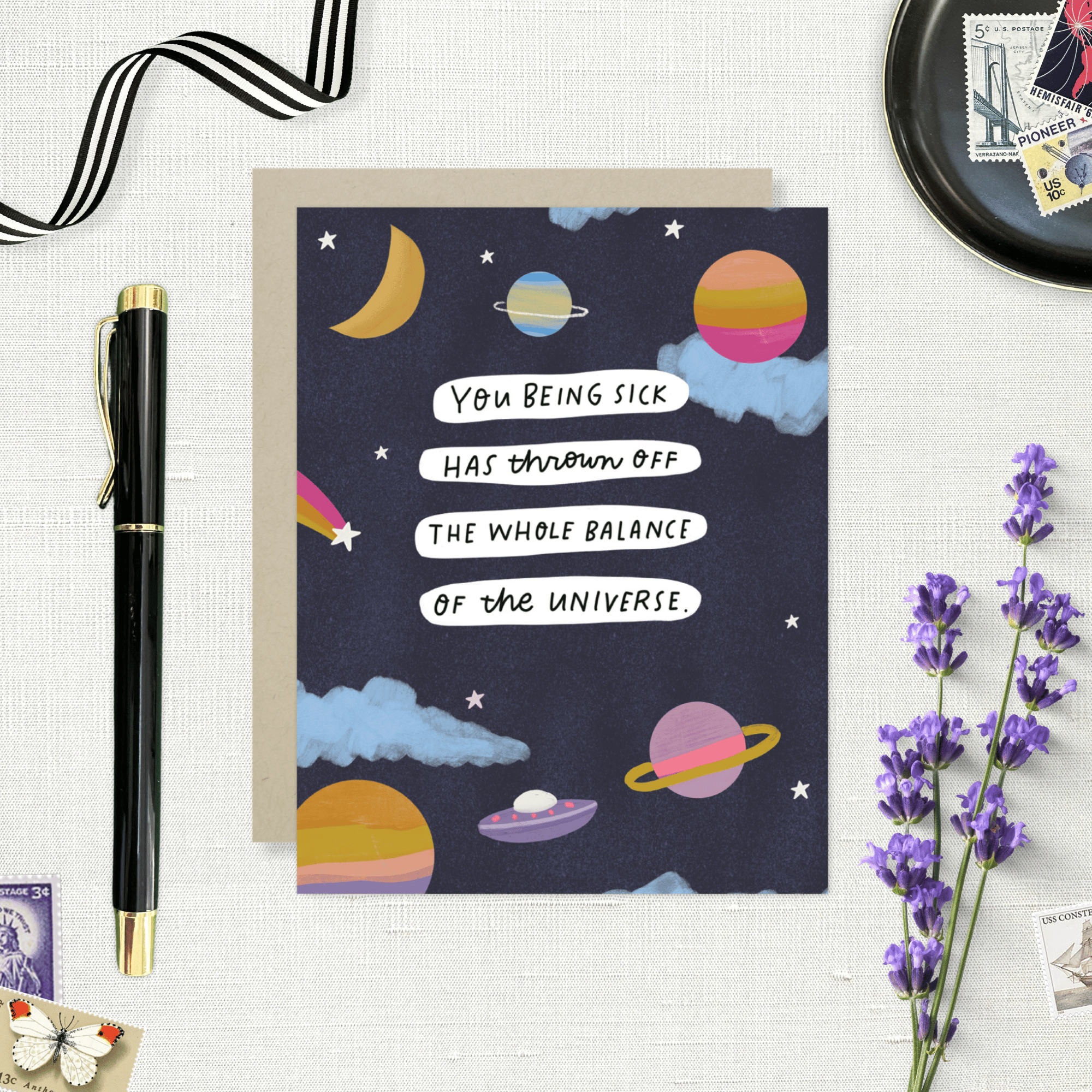Get well greeting card with space-themed design and humorous text on a white surface with a pen and lavender flowers.