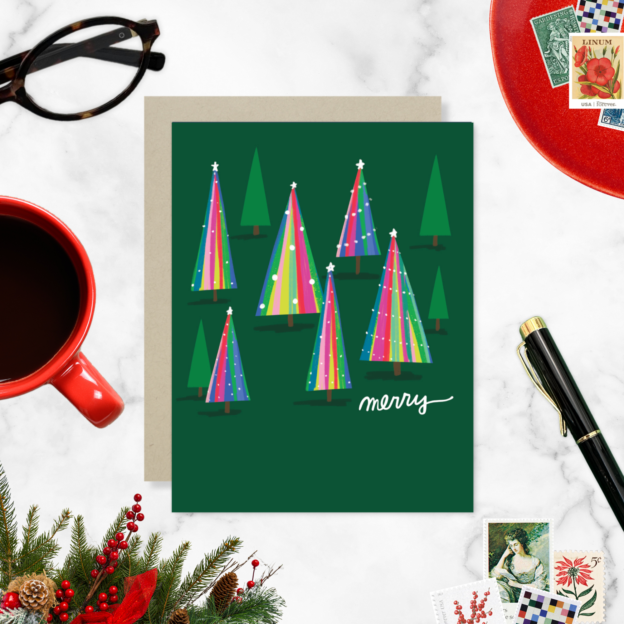 Christmas greeting card with colorful trees and 'merry' text on a white surface with a cup of coffee and decorative items.