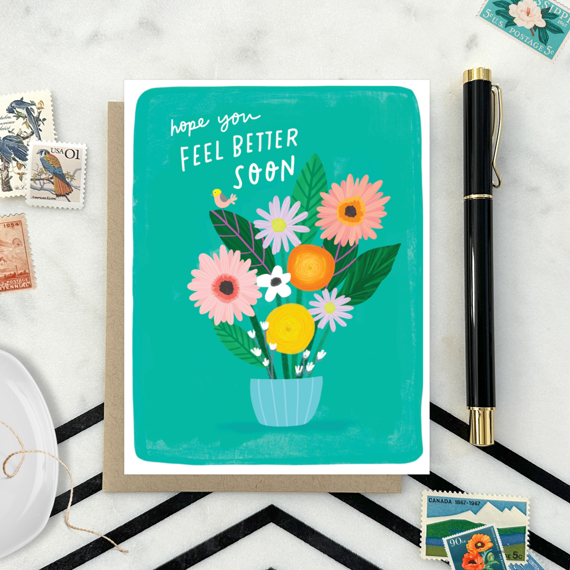 Get well greeting card with floral design and text 'hope you feel better soon' on a marble surface with stamps and a pen.