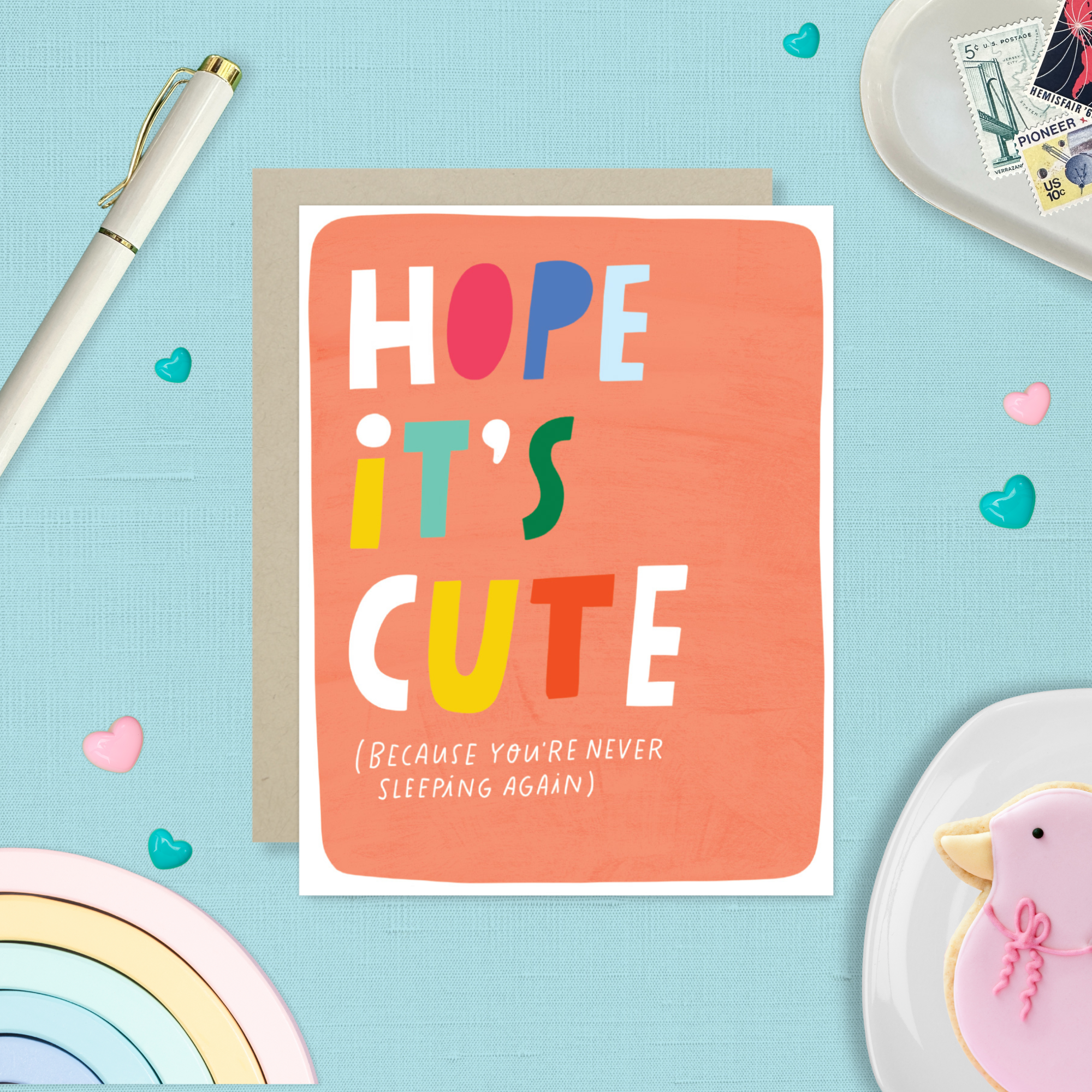 New baby greeting card colorful with colorful words 'Hope it's cute' text on a light blue background with decorative items.