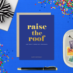A birthday greeting card with 'raise the roof' printed in gold foil. Below it, in smaller gold foil text, reads 'Just don’t throw out your back.