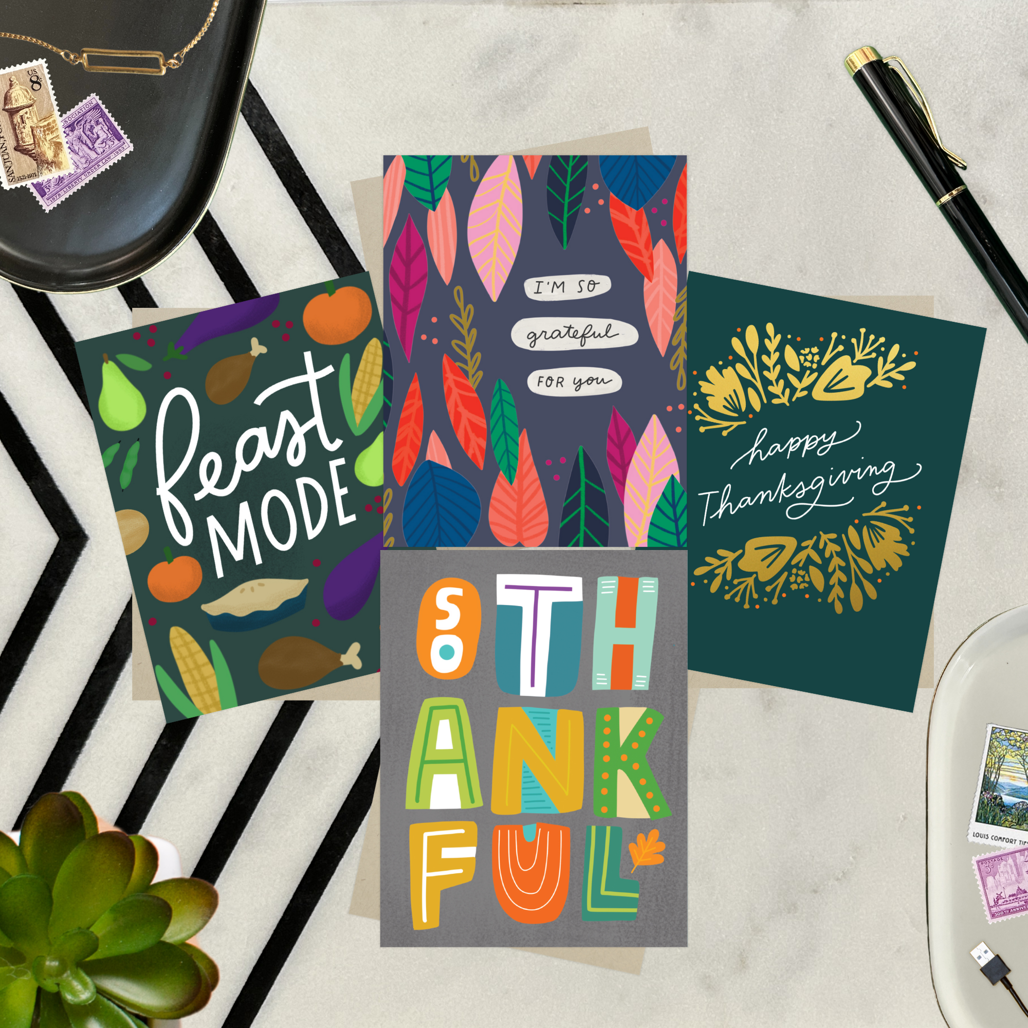 Four colorful greeting cards with autumn-themed designs on a marble surface featuring "I'm so grateful for you," "Happy Thanksgiving," "Feast Mode," and "So Thankful."