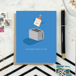 A playful birthday greeting card with an illustration of a smiling slice of toast with a party hat popping out of a toaster, and the text 'A Birthday Toast to You!'.