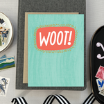 A congratulations greeting card with a hand-lettered word 'Woot!' written in bold, fun typography on a teal background with a textured appearance.