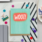A congratulations greeting card with a hand-lettered word 'Woot!' written in bold, fun typography on a teal background with a textured appearance.