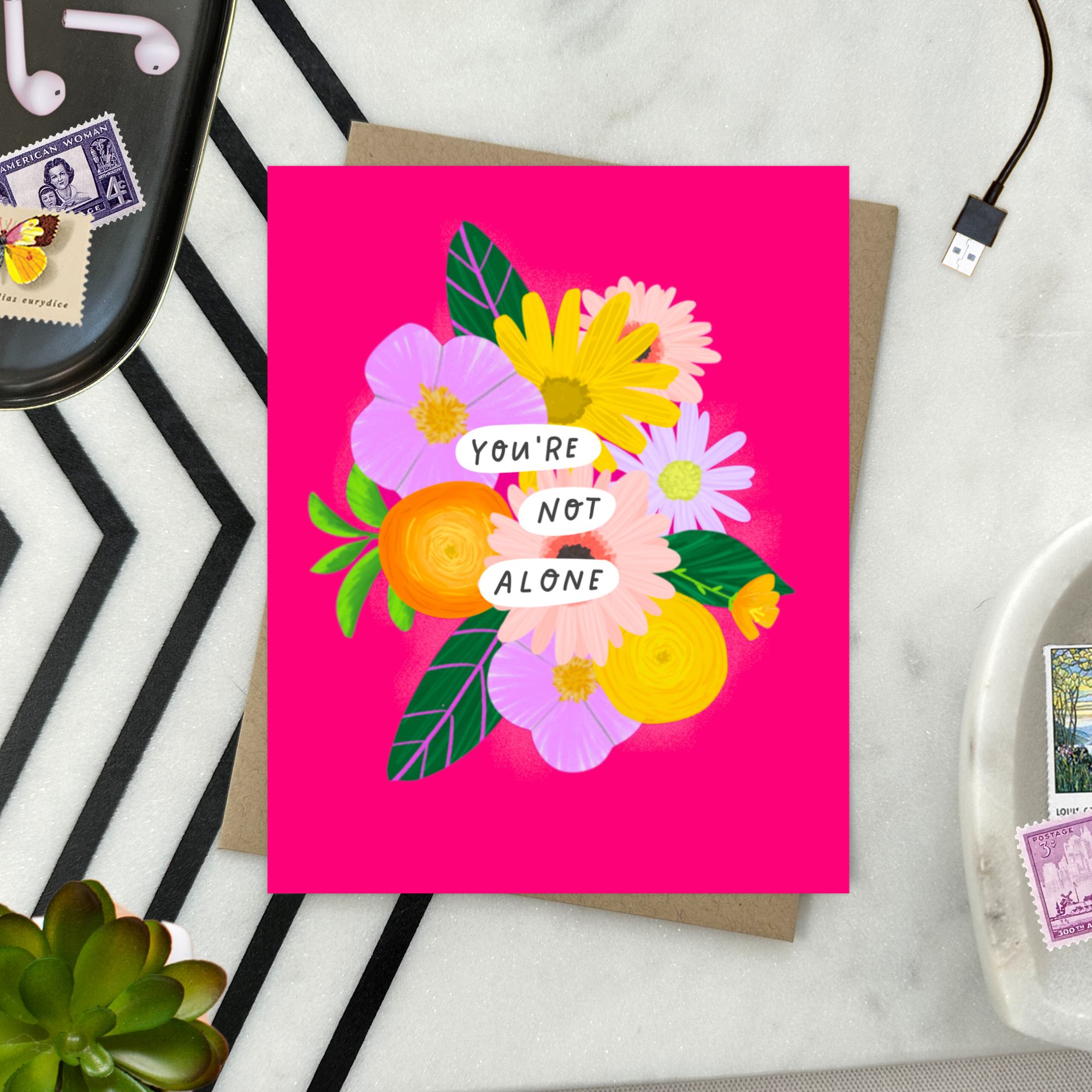 Colorful friendship or encouragement greeting card featuring a modern floral bouquet in pinks, oranges, yellows, and lavenders with the handwritten words, "You Are Not Alone" on a hot pink background. 